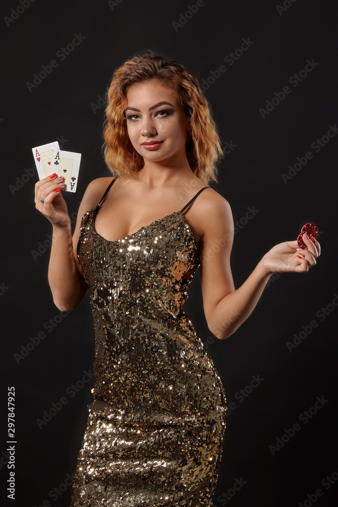 Ginger girl in shiny dress posing holding two playing cards and chips in her hands standing against black studio background. Casino, poker. Close-up. - obrazy, fototapety, plakaty 