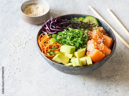 Hawaiian salmon poke bowl with rice, avocado, cucumber, carrot, red cabbage, chukka and sesame seeds on a gray background.