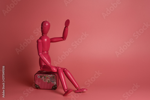 A pink wooden man sits on a suitcase against a pink background. Isolated. Business, selling out, late, waiting, flying concept. Copy space, banner, postcard.