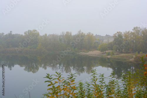 Beautiful landscape, autumnal nature with white dense fog over Career Lake, Dnipro city, Ukraine. Tall trees and large shrubs with yellow leaves around a clean and beautiful lake.