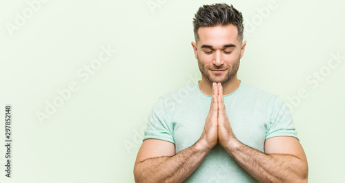 Young handsome man against a green background holding hands in pray near mouth, feels confident. photo