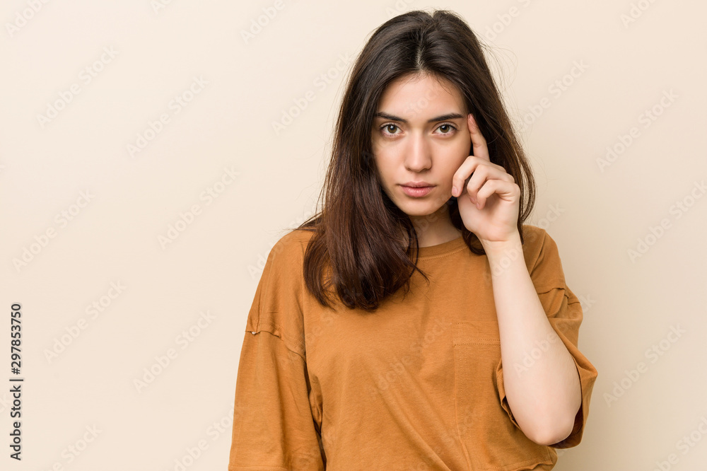 Young brunette woman against a beige background pointing temple with finger, thinking, focused on a task. Young brunette woman against a beige background pointing temple with finger, thinking, focused on a task.
