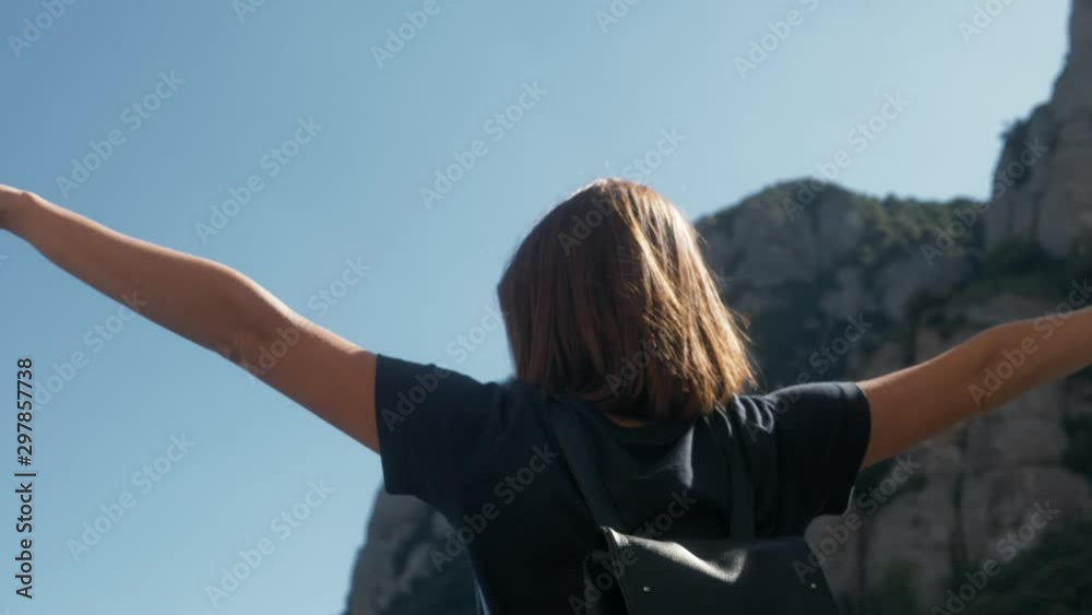 Young woman tourist with backpack standing on cliff's edge top mountain with raised hands and looking to a sky Young woman tourist with backpack standing on cliff's edge top mountain with raised hands and looking to a sky