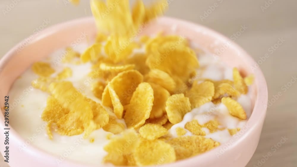 Dry breakfast cereal cornflakes falling in pink bowl with milk in slowmotion on wooden background. Food close up. Dry breakfast cereal cornflakes falling in pink bowl with milk in slowmotion on wooden background. Food close up.