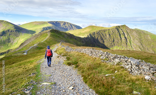A hiker walking along a stone chip path on a mountain ridge towards the summits of Hobcarton and Hopegill Head in the Lake District, England, UK.