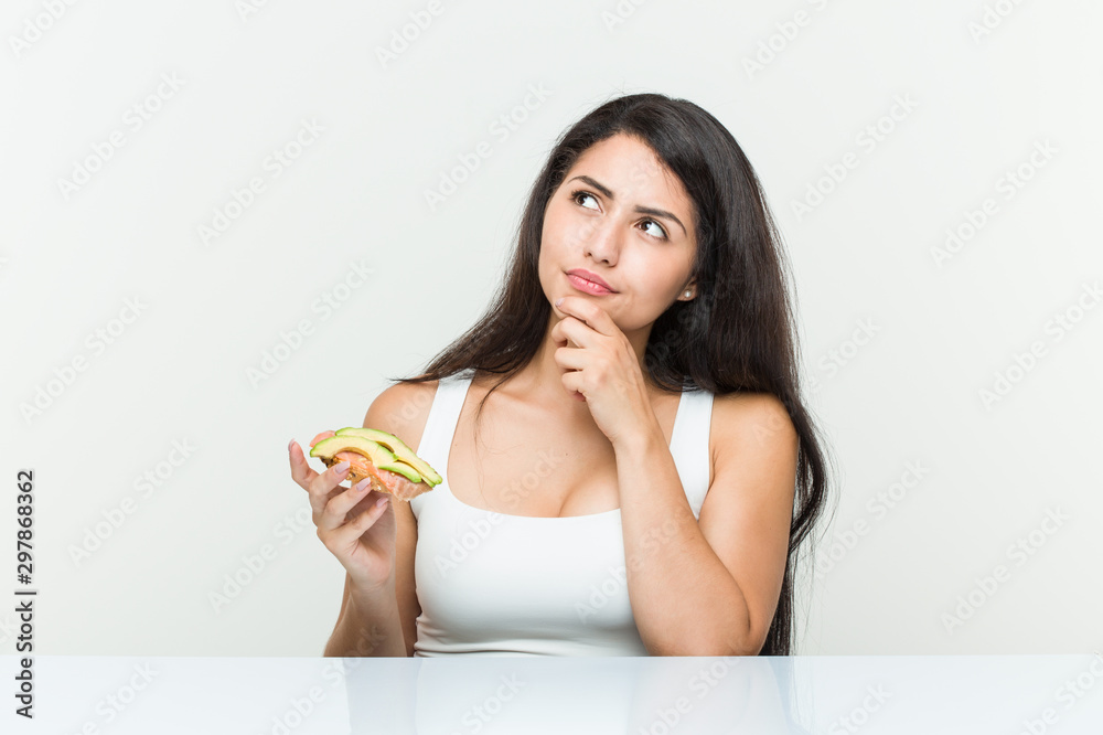 Young hispanic woman holding an avocado toast looking sideways with doubtful and skeptical expression. Young hispanic woman holding an avocado toast looking sideways with doubtful and skeptical expression.