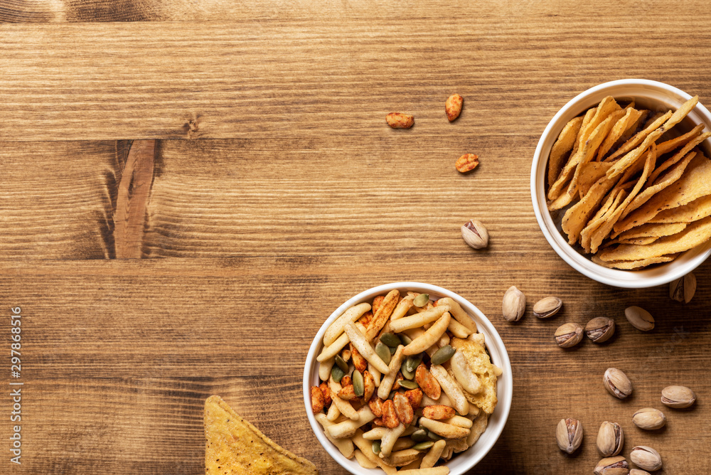Various beer snacks in white bowls on a wooden table, top view, flat lay. Nachos, pistachios, cheese sticks and other beer snacks on a wooden background, free space for text. Various beer snacks in white bowls on a wooden table, top view, flat lay. Nachos, pistachios, cheese sticks and other beer snacks on a wooden background, free space for text.