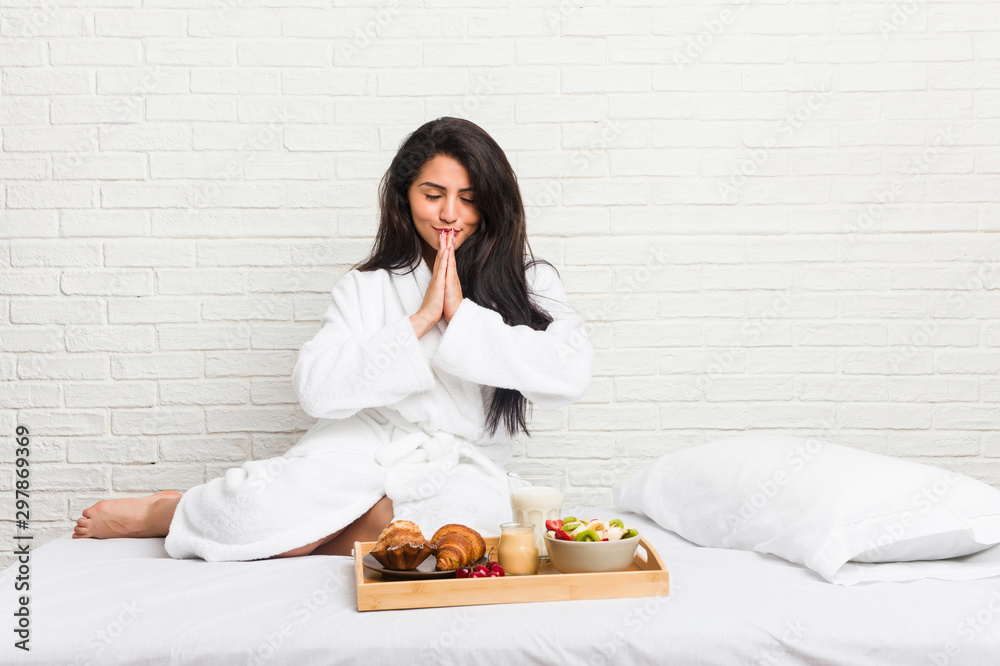 Young curvy woman taking a breakfast on the bed holding hands in pray near mouth, feels confident.