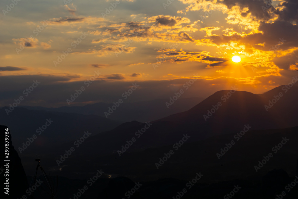 Sun setting behind dramatic clouds and beyond dark silhouette mountains