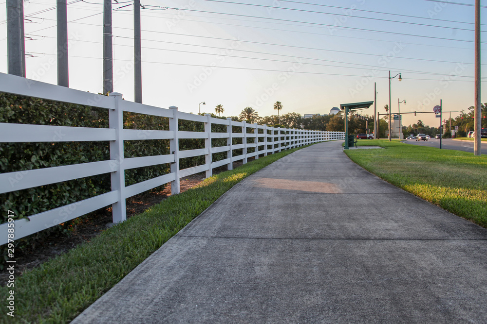 A long, curved white wooden fence alongside a grassy walk in the background of the beautiful landscape of plants and trees in the late afternoon in Celebration, Orlando, Florida, USA.