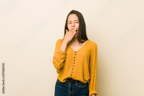 Young hispanic woman against a beige background yawning showing a tired gesture covering mouth with hand.