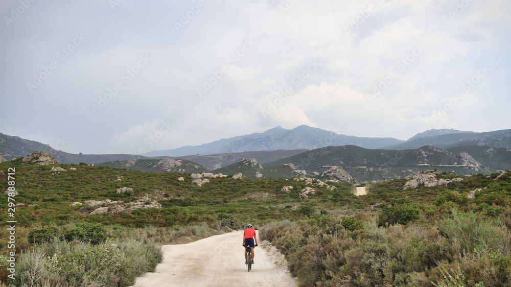Cycling. Offroad Cycling in Agriates Desert, Corsica, France. a red shirt cyclist, Dusty road, Rocky road, Mediterranean Vegetation. Mountains, Daylight. Cloudy sky. Courage for the Journey. 