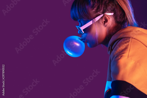 Teen igen girl wear stylish trendy sunglasses and hoodie blowing bubble gum profile side view, pretty young woman fashion cool model with bubblegum 80s at party purple studio background, copy space