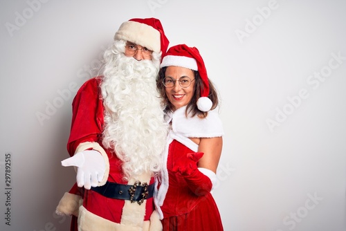 Middle age couple wearing Santa costume hugging over isolated white background smiling cheerful offering palm hand giving assistance and acceptance.