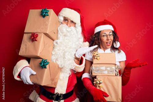 Middle age couple wearing Santa costume holding tower of gifts over isolated red background pointing aside with hands open palms showing copy space, presenting advertisement smiling excited happy photo