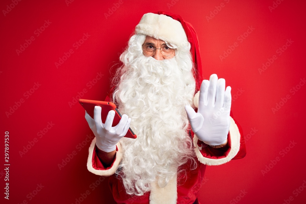Middle age man wearing Santa Claus costume using tablet over isolated red background with open hand doing stop sign with serious and confident expression, defense gesture