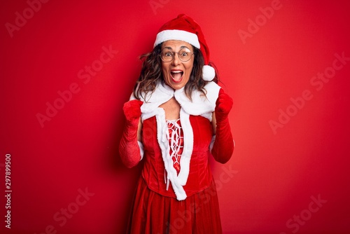 Middle age beautiful woman wearing Santa Claus costume over isolated red background celebrating surprised and amazed for success with arms raised and open eyes. Winner concept.