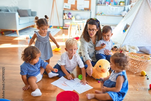 Beautiful teacher and group of toddlers sitting on the floor drawing using paper and pencil around lots of toys at kindergarten photo