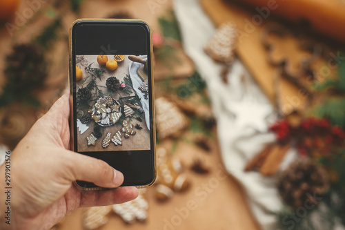 Hand holding phone and taking photo of christmas composition of festive gingerbread cookies, anise, cinnamon, pine cones, cedar branches on rustic table. Merry Christmas. Holiday workshop