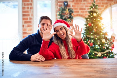 Young couple wearing santa claus hat sitting on chair and table around christmas tree at home showing and pointing up with fingers number ten while smiling confident and happy.