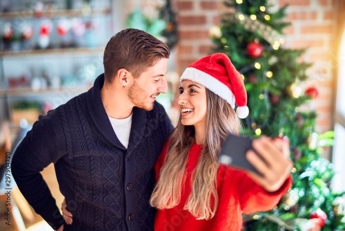Young beautiful couple smiling happy and confident. Standing and hugging make selfie by camera around christmas tree at home