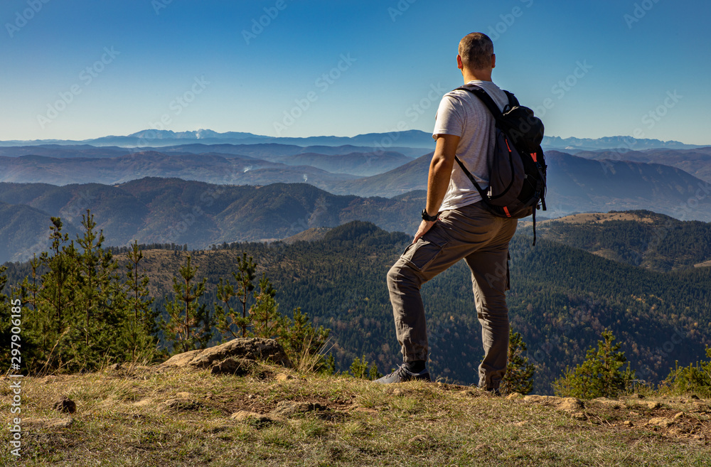 Naklejka premium Rear view of male hiker with backpack standing on top of the mountain and enjoying the view during the day.