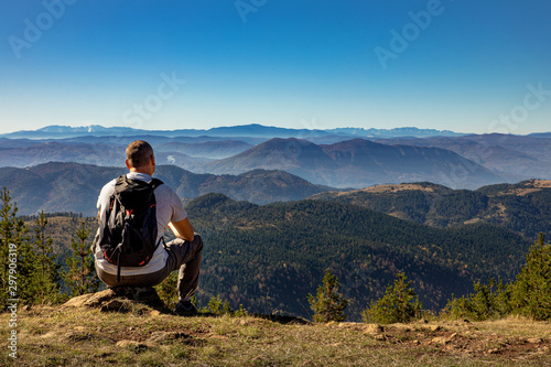 Rear view of male hiker with backpack sitting on top of the mountain and enjoying the view during the day.