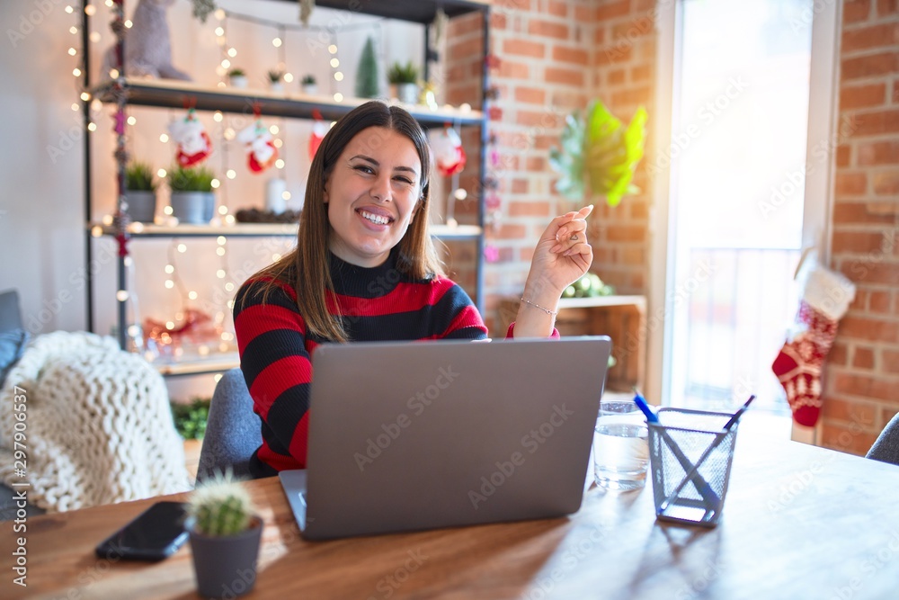 Beautiful woman sitting at the table working with laptop at home around christmas lights with a big smile on face, pointing with hand and finger to the side looking at the camera. Beautiful woman sitting at the table working with laptop at home around christmas lights with a big smile on face, pointing with hand and finger to the side looking at the camera.