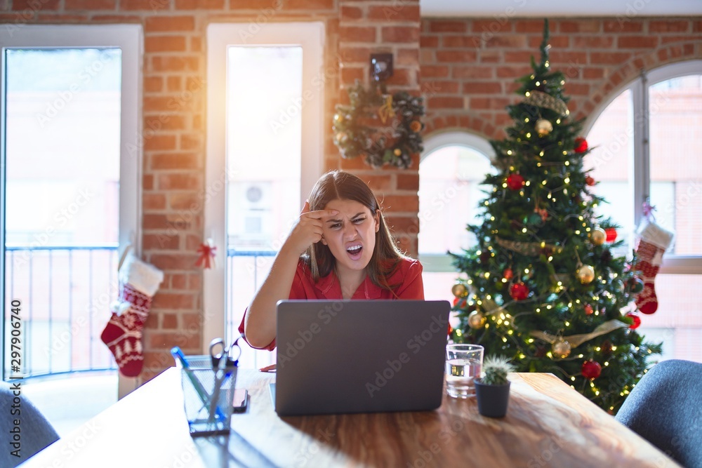 Beautiful woman sitting at the table working with laptop at home around christmas tree pointing unhappy to pimple on forehead, ugly infection of blackhead. Acne and skin problem Beautiful woman sitting at the table working with laptop at home around christmas tree pointing unhappy to pimple on forehead, ugly infection of blackhead. Acne and skin problem