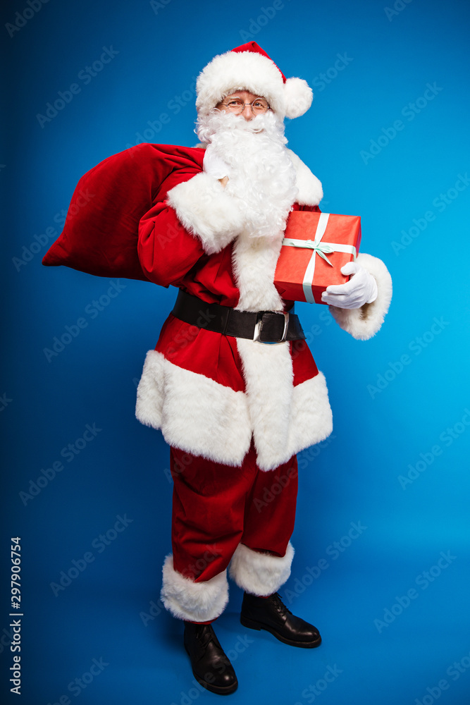 Happy Santa. Full-length photo of Santa Claus, posing with a sack of presents on his right shoulder and a present in a red wrapping paper in his right arm, looking at the camera. Happy Santa. Full-length photo of Santa Claus, posing with a sack of presents on his right shoulder and a present in a red wrapping paper in his right arm, looking at the camera.