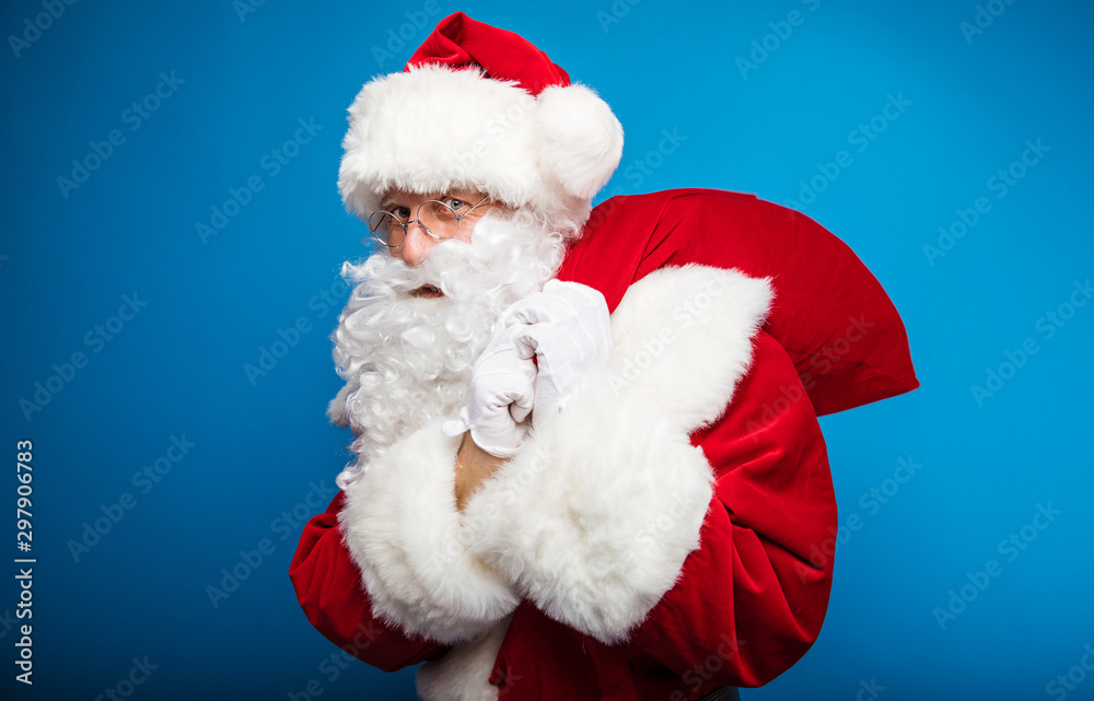 Santa is sneaking. Close up photo of Santa, who is posing in profile, holding a sack with presents on his left shoulder with both of his hands and looking at the camera.