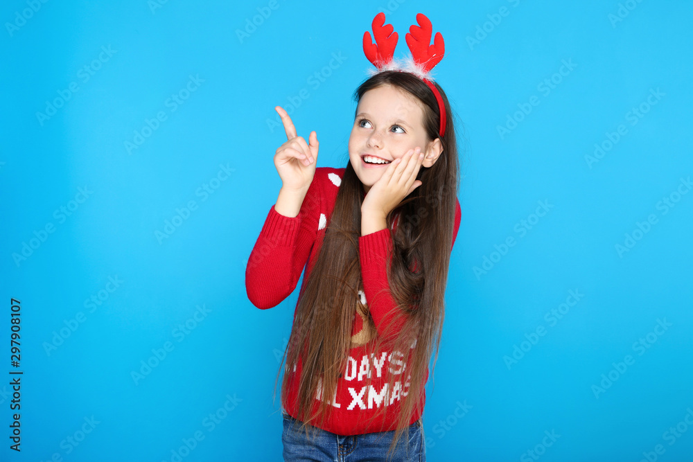Little girl in christmas sweater and red horns pointing by finger on blue background Little girl in christmas sweater and red horns pointing by finger on blue background