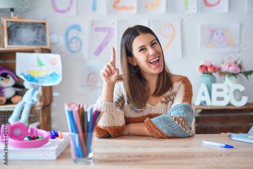Young beautiful teacher woman wearing sweater and glasses sitting on desk at kindergarten pointing finger up with successful idea. Exited and happy. Number one.