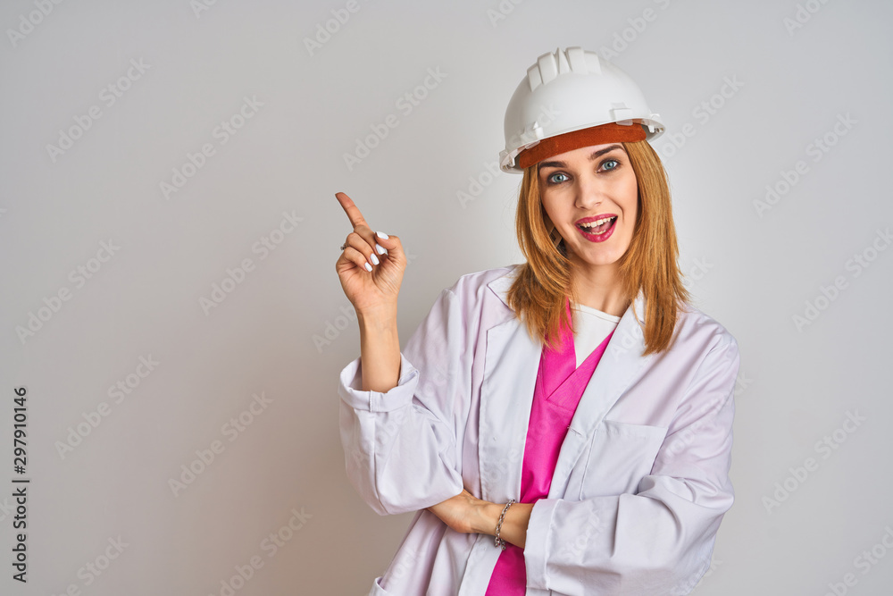 Redhead caucasian woman engineer wearing safety helmet over isolated background with a big smile on face, pointing with hand and finger to the side looking at the camera. Redhead caucasian woman engineer wearing safety helmet over isolated background with a big smile on face, pointing with hand and finger to the side looking at the camera.