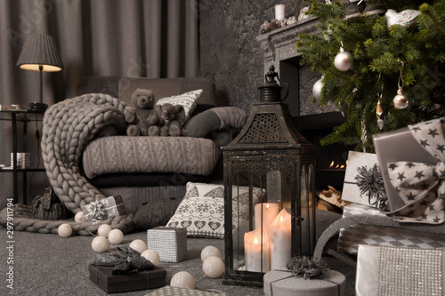 Modern cozy interior in gray shades with an armchair in front of fireplace and decorated Christmas tree. On the chair there are nice knitted blanket, cushions and Teddy bear. 