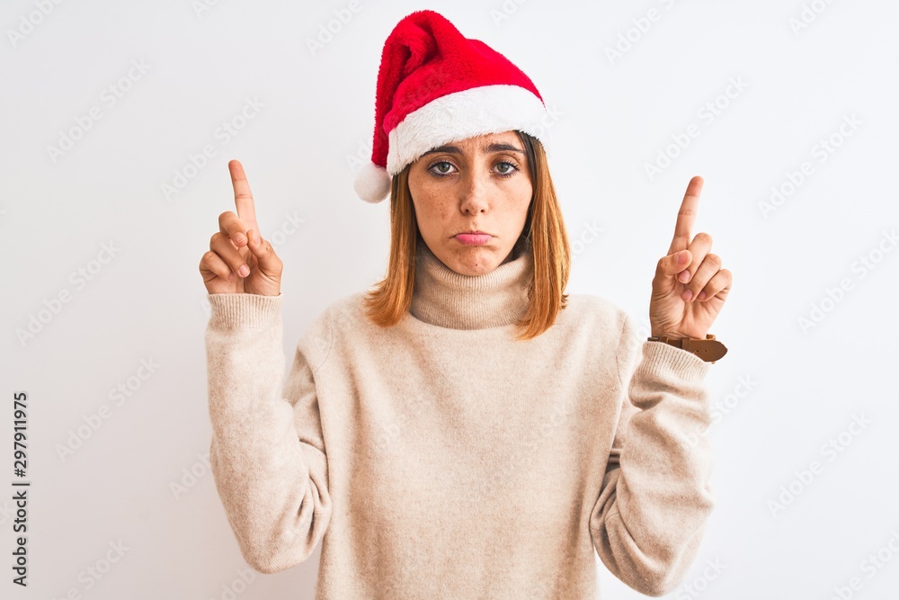 Beautiful redhead woman wearing christmas hat over isolated background Pointing up looking sad and upset, indicating direction with fingers, unhappy and depressed. Beautiful redhead woman wearing christmas hat over isolated background Pointing up looking sad and upset, indicating direction with fingers, unhappy and depressed.