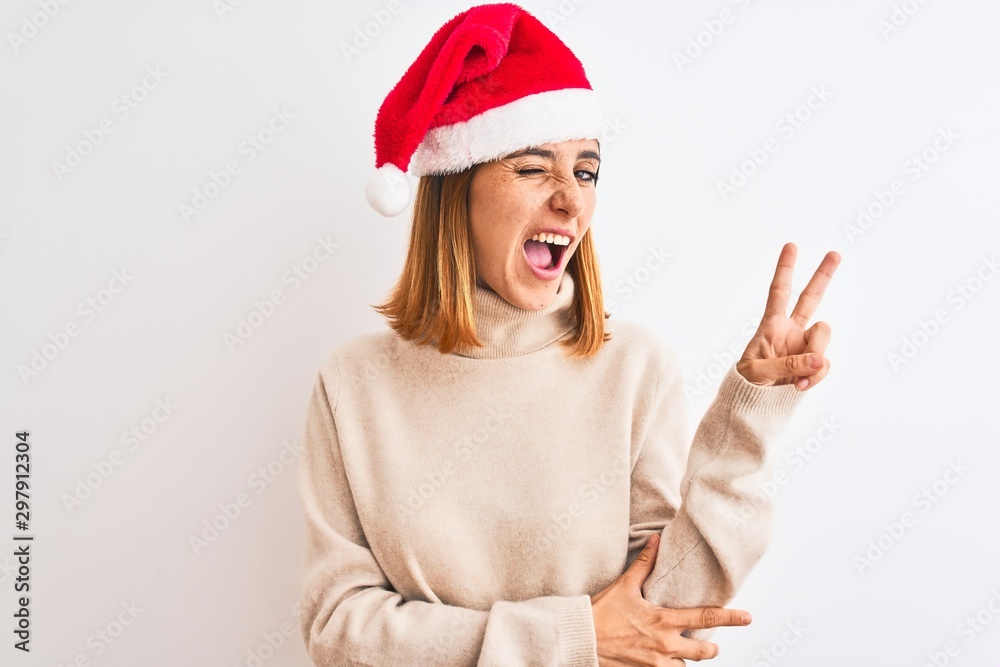 Beautiful redhead woman wearing christmas hat over isolated background smiling with happy face winking at the camera doing victory sign with fingers. Number two. Beautiful redhead woman wearing christmas hat over isolated background smiling with happy face winking at the camera doing victory sign with fingers. Number two.