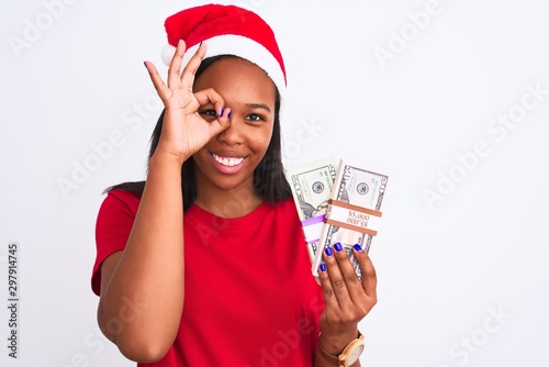 Young african american woman wearing christmas hat and holding bunch of dollars banknotes with happy face smiling doing ok sign with hand on eye looking through fingers © Krakenimages.com