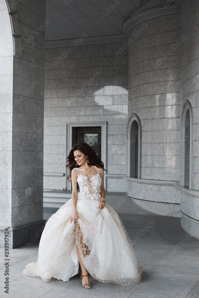 Fototapeta premium A happy young bride with a wedding hairstyle in a white lace dress runs through the courtyard of an ancient church. Beautiful model girl holds her wedding dress in her hands and poses in the old city