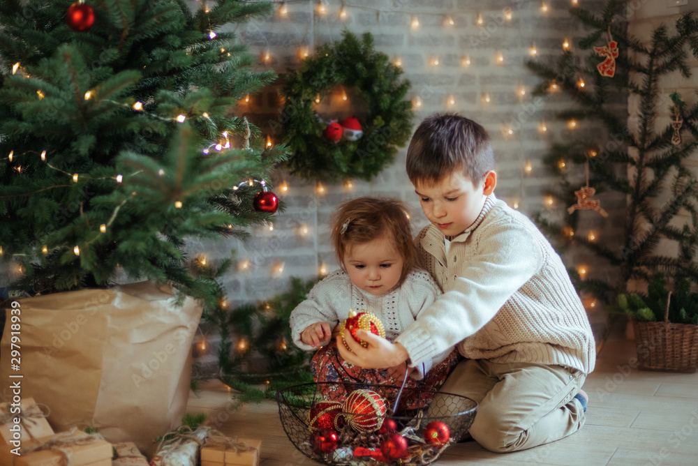 Brother and little sister at the Christmas tree decorate with Christmas decor. New Year happy family concept. Christmas Miracle Brother and little sister at the Christmas tree decorate with Christmas decor. New Year happy family concept. Christmas Miracle