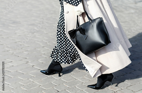 Woman in autumn or spring in a beige coat and a black dress with white polka dots. Fashionable bag close-up in female hands.Girl walks in the city outdoors. Stylish modern and feminine image, style.