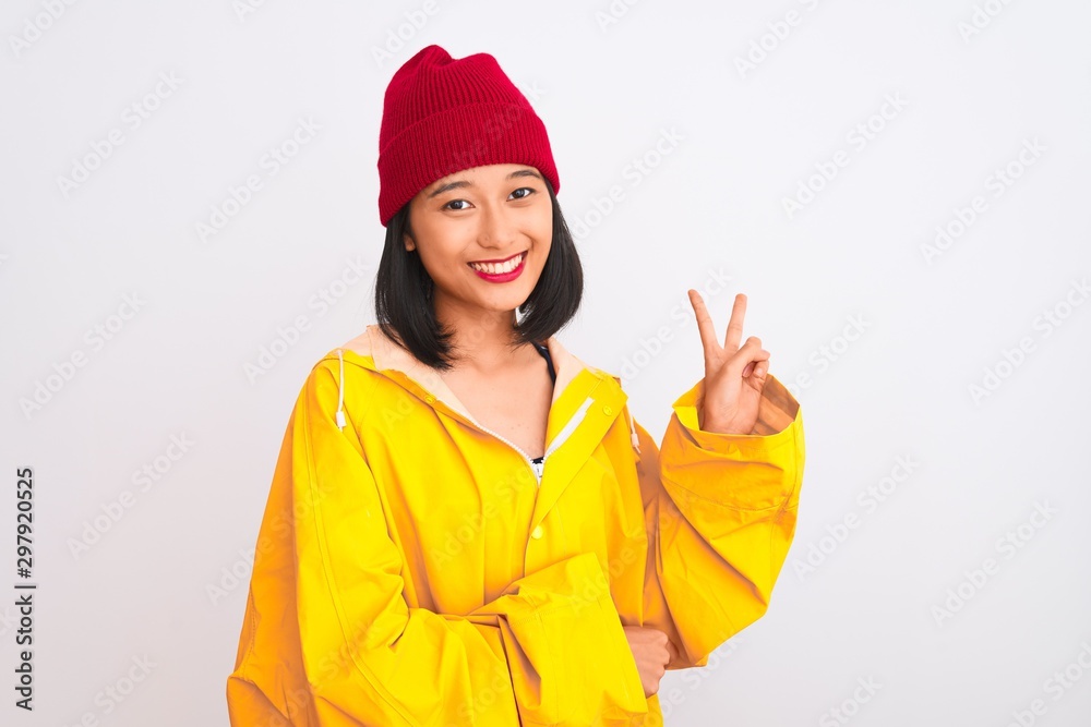 Young beautiful chinese woman wearing raincoat and wool cap over isolated white background smiling with happy face winking at the camera doing victory sign. Number two. Young beautiful chinese woman wearing raincoat and wool cap over isolated white background smiling with happy face winking at the camera doing victory sign. Number two.