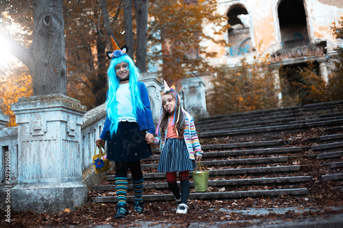 Two cheerful little girls, in colorful halloween costume of unicorn, blue hair and green bucket for sweets, standing on stairs behind scary abandoned house. © Lalandrew