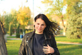white girl with long dark hair stands in a leather jacket on a background of autumn yellow trees and grass with fallen leaves