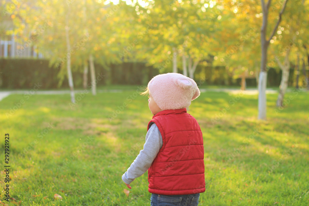 A small child in a fluffy pink hat and red vest is running in the autumn park. Beautiful fall sunny day outdoors. A small child in a fluffy pink hat and red vest is running in the autumn park. Beautiful fall sunny day outdoors.