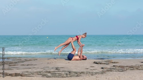 Muscular man and fit young woman training together in Acro Yoga. Morning sun lighting the couple and the mediterranean beach background.