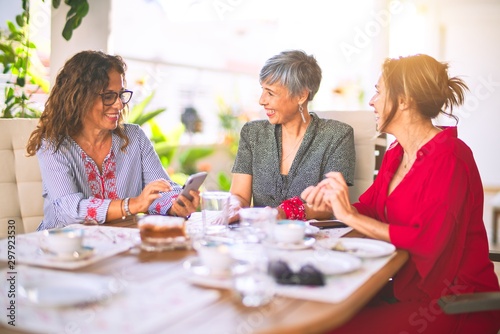 Meeting of middle age women having lunch and drinking coffee. Mature friends smiling happy using smartphone at home on a sunny day