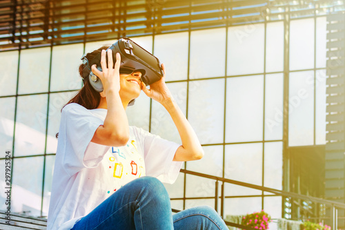 Young female with long hair sitting on the stairs and wearing virtual reality glasses with big headphones. Wide smile and hands holding the device. Glass business building in the background.