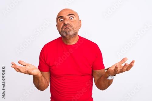 Middle age handsome man wearing red casual t-shirt standing over isolated white background clueless and confused expression with arms and hands raised. Doubt concept.