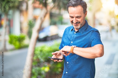 Middle age handsome businessman standing on the street using smartphone looking at the watch